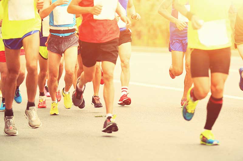 A group of runners wearing white race bibs on various brightly-colored t-shirts, all wearing brightly colored shorts, socks and running shoes. They are running toward the camera.