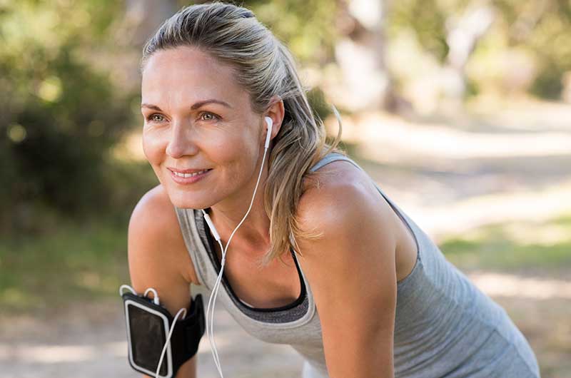 Smiling white woman in her 30s wearing a gray tank top is listening to music on her earbuds that are connected to her phone. Her phone is strapped to her upper right arm. SHe appears to be taking a break from exercising by bending over and resting her hand on her upper legs, but below the waist is not shown.