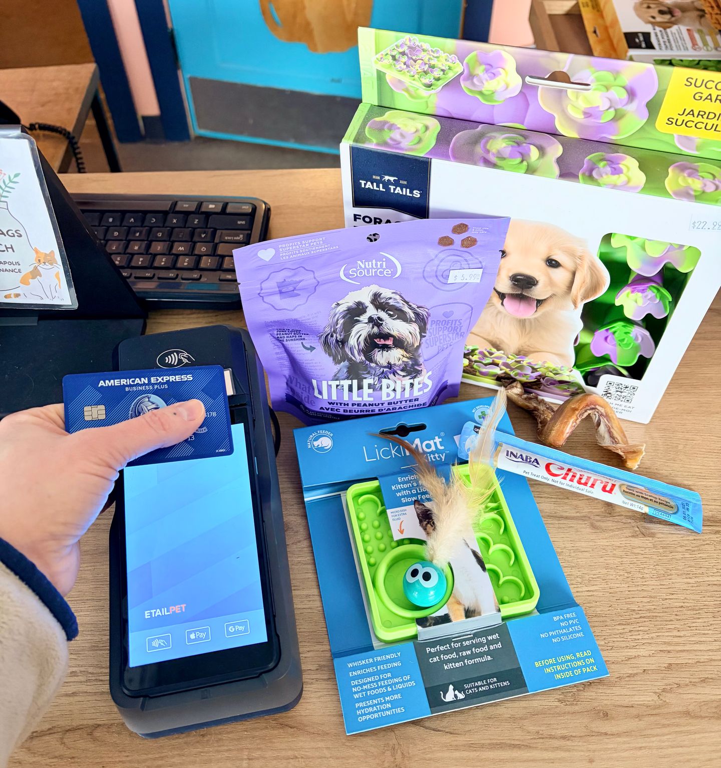 person holds a credit card to a card reater at a checkout counter. An assortment of pet treats and toys are on the counter