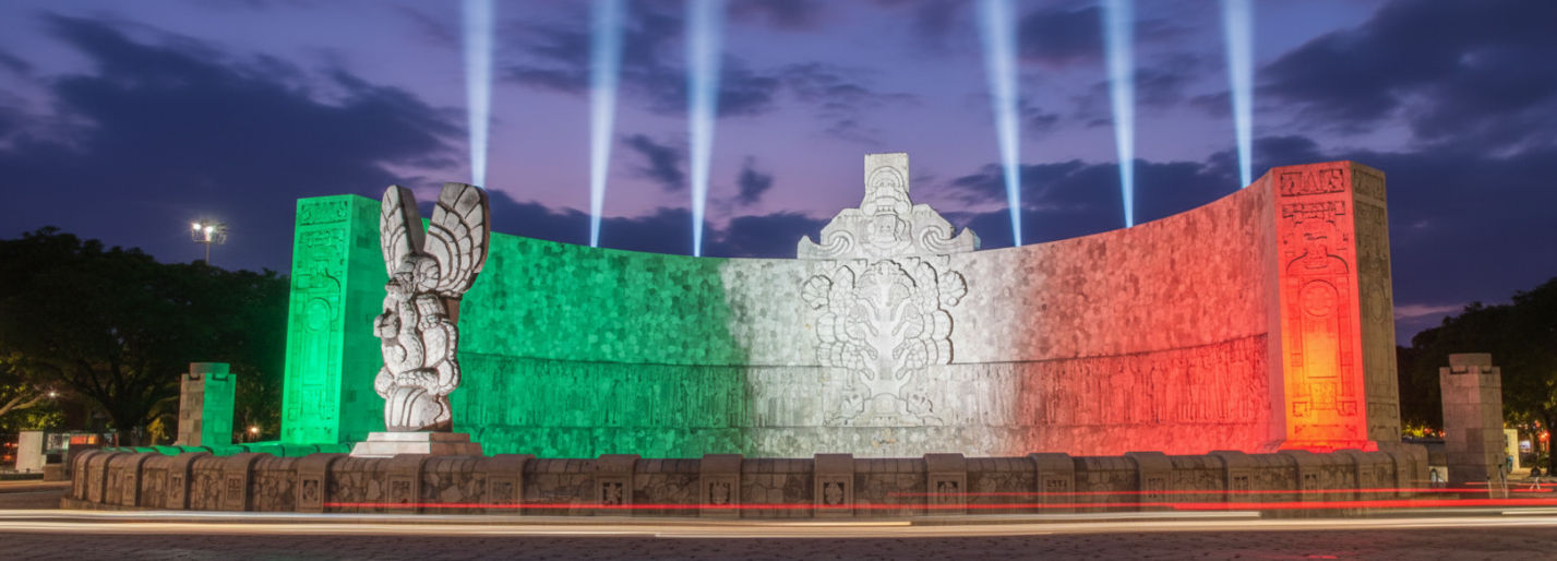 Una vista frontal y amplia del Monumento a la Patria en Mérida, bajo un cielo despejado, con la bandera mexicana ondeando y la majestuosidad de la estructura resaltando.