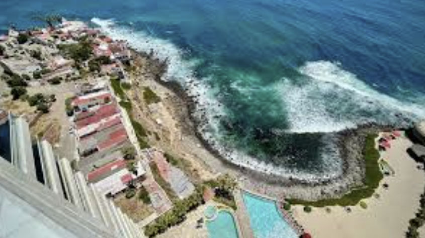 Aerial view of a coastal resort featuring bright blue swimming pools, red umbrellas, and a cluster of red-roofed buildings next to a rocky shoreline with crashing ocean waves.