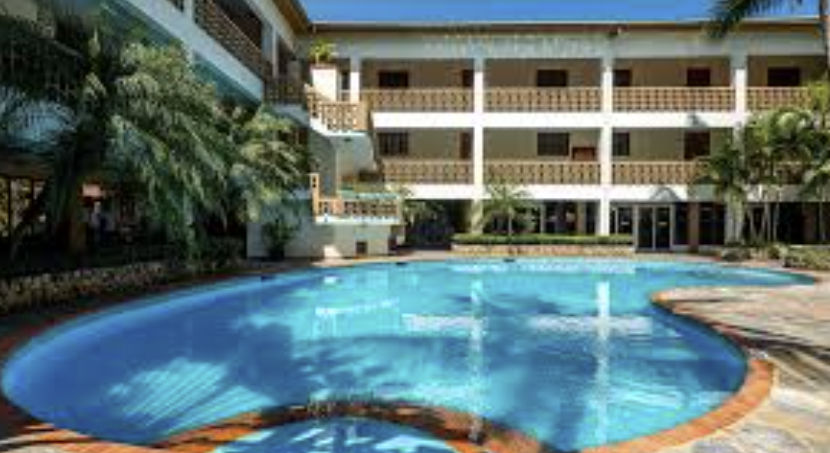Large outdoor swimming pool at a resort, surrounded by palm trees and a white three-story building with balconies