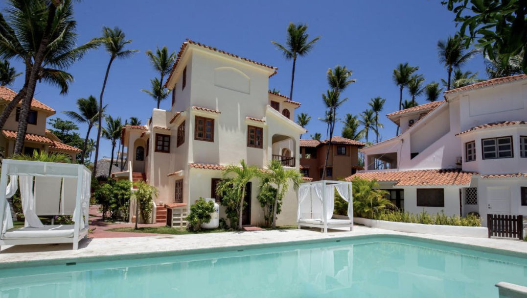 View of yellow Mediterranean-style villas surrounded by palm trees facing the white sand beach and turquoise waters at Los Corales Beach Village, Punta Cana.