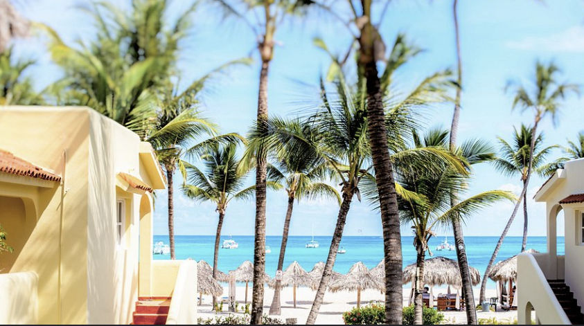 View of yellow Mediterranean-style villas surrounded by palm trees facing the white sand beach and turquoise waters at Los Corales Beach Village, Punta Cana.