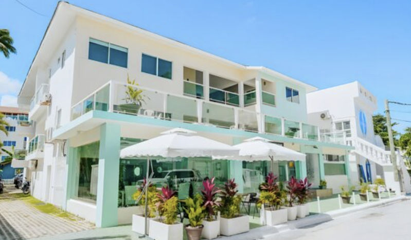 Una vista exterior de la fachada del Green Coast Beach Hotel en Punta Cana, que muestra un edificio moderno de tres pisos pintado de blanco y verde menta. La planta baja cuenta con una terraza de cafetería al aire libre con sombrillas blancas, mesas y gra