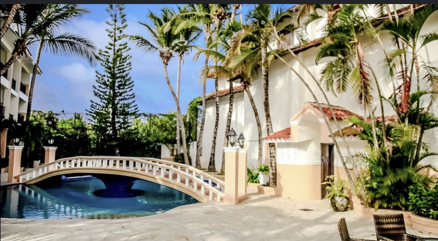 Exterior view of a tropical hotel courtyard featuring a deep blue swimming pool and an arched stone bridge with a white railing. The pool area is surrounded by textured stone paving, numerous tall palm trees, and a large central pine tree, all under a par
