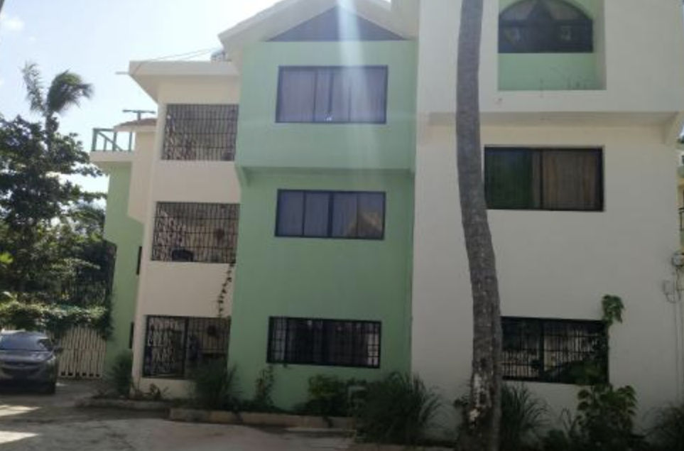 Exterior view of the three-story building at Riviera Punta Cana Eco Travelers in Bavaro, Dominican Republic, showing its light green and white facade with dark windows and tropical greenery around it.