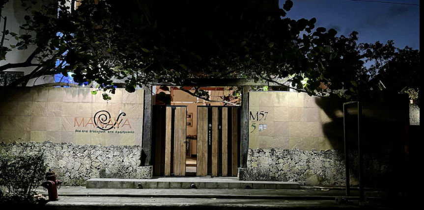 Fachada exterior iluminada de noche de Manaya Bed and Breakfast and Apartments en Punta Cana, con un muro de piedra y baldosas, portón de madera y árboles cubriendo la entrada.