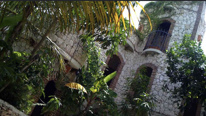 Rustic light stone building with brick arches and a balcony, surrounded by lush tropical vegetation with palm trees and vines.