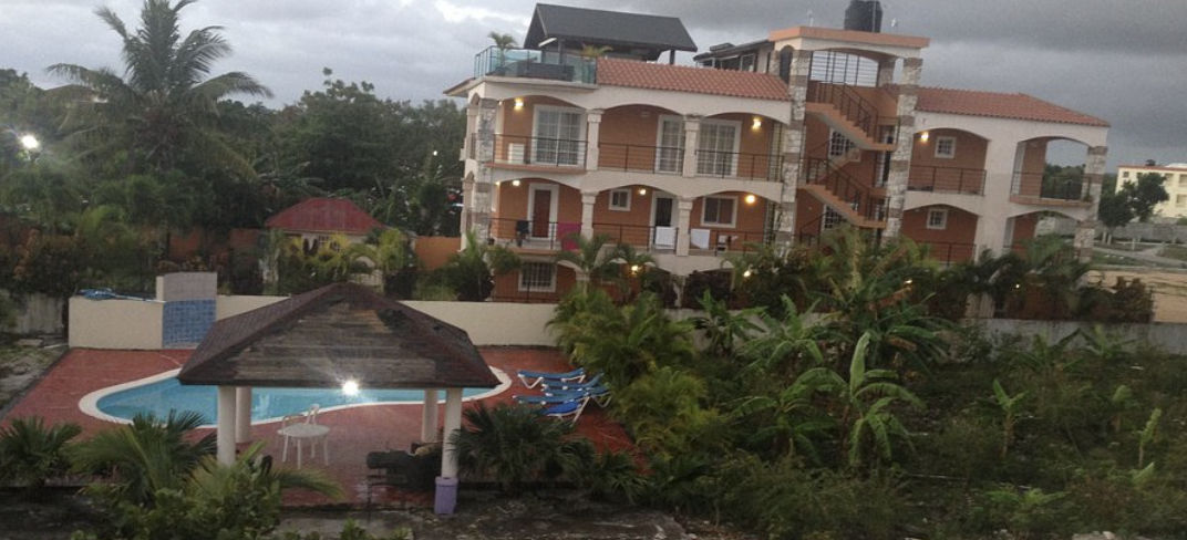 Una vista elevada de un complejo de apartamentos de tres pisos con balcones arqueados y una escalera de piedra exterior, situado detrás de una exuberante zona de piscina y jardín tropical. En el primer plano, hay una piscina en forma de riñón, un cenador 