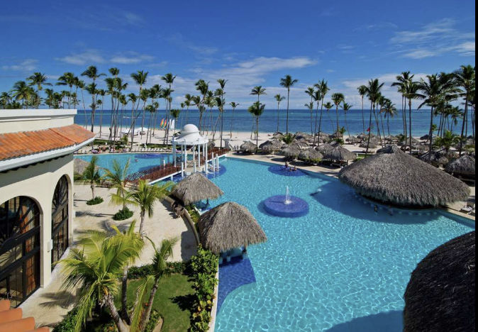Aerial view of a luxurious tropical resort with a large swimming pool and palm trees, located next to a white sand beach and the blue ocean under a clear sky.