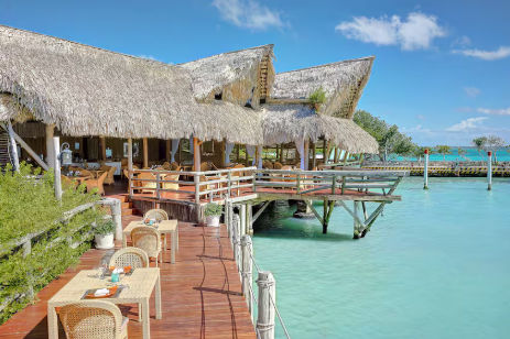 Restaurante de estilo tropical con techos de palapa y terraza de madera sobre aguas cristalinas color turquesa, con mesas de mimbre preparadas para comer al aire libre.