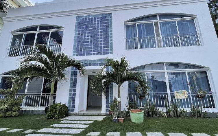 Fachada blanca de dos pisos del Hostal Gava Punta Cana con ventanas grandes, detalles de bloques de vidrio azul y palmeras en el jardín delantero.