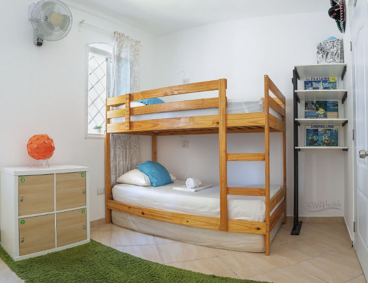 Fachada blanca de dos pisos del Hostal Gava Punta Cana con ventanas grandes, detalles de bloques de vidrio azul y palmeras en el jardin delantero