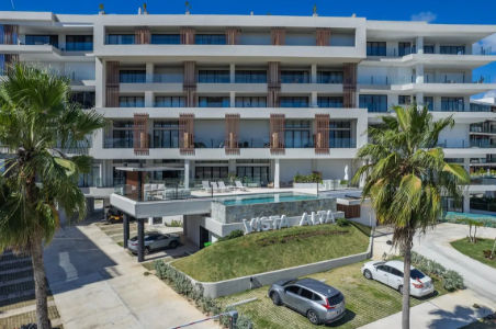 Modern facade of the Vista Alta residential complex in Cap Cana, Punta Cana, showing the elevated swimming pool, palm trees, and outdoor parking area under a clear blue sky.
