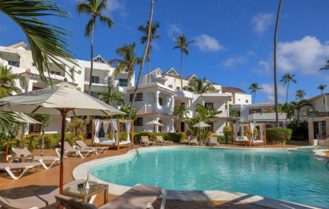 Outdoor hotel swimming pool surrounded by lounge chairs, umbrellas, cabana beds, and palm trees, with white buildings in the background under a blue sky.