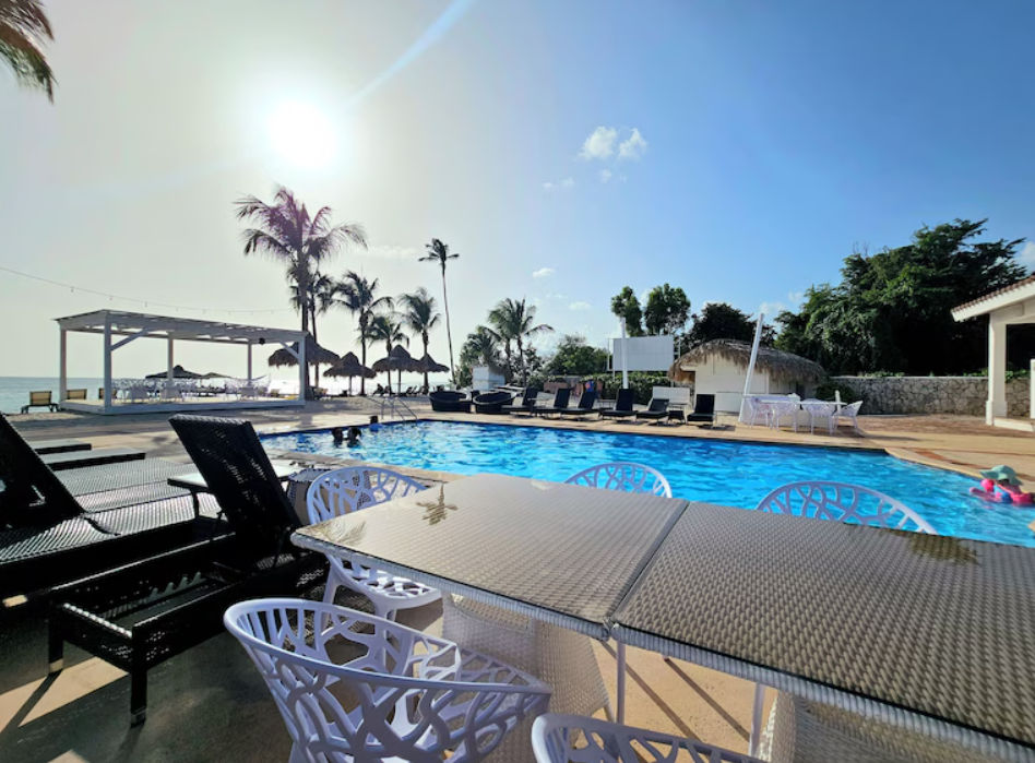 Piscina al aire libre con camastros y mesas junto a la playa en un resort tropical de Punta Cana bajo un cielo soleado