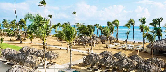 Panoramic view of a luxury beach resort with swaying palm trees, palapa umbrellas, lounge chairs on the golden sand, and the turquoise Caribbean Sea in the background