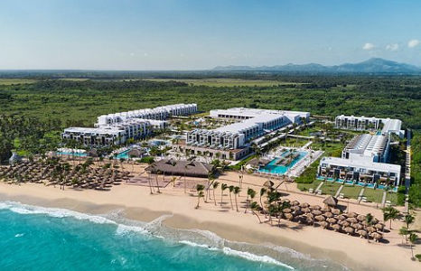 Panoramic aerial view of a luxury beachfront resort with modern white buildings, pools, and terraces. A light sand beach with palm trees and thatched-roof cabanas borders the turquoise sea. A green jungle and mountains stretch into the background under a 
