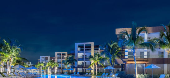 Night view of the main illuminated pool at the Radisson Blu Resort & Residence Punta Cana, surrounded by palm trees, umbrellas, and modern hotel buildings.