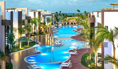 Night view of the main illuminated pool at the Radisson Blu Resort & Residence Punta Cana, surrounded by palm trees, umbrellas, and modern hotel buildings.