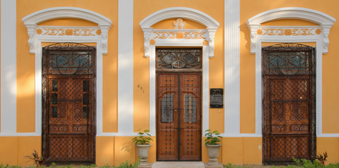 Fotografía de una hermosa casa de estilo colonial moderno en un vecindario frondoso y tranquilo cerca del estadio. La luz del sol se filtra a través de los árboles, evocando una sensación de paz, lujo y hogar, representativo del catálogo de Itzimná Real Estate.
