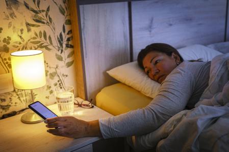 Close up of mature woman laying on bed reaching her mobile phone on the night table early in the morning. Selective focus on hand and clock. High resolution 42Mp indoors digital capture taken with SONY A7rII and Zeiss Batis 40mm F2.0 CF lens