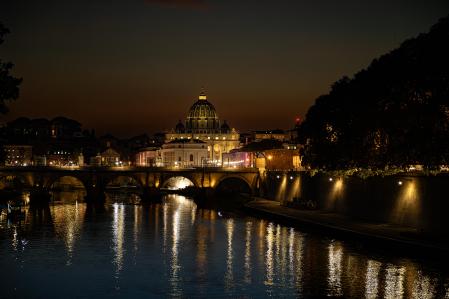 La majestuosa cúpula de la Basílica de San Pedro, iluminada y dominando el horizonte, mientras que el puente que cruza el Tíber se refleja en las aguas tranquilas del río.