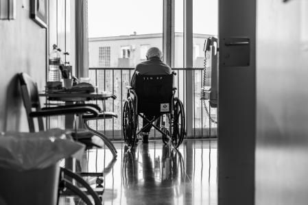 Un anciano mira por la ventana de un hospital, indiferente a las actividades navideñas que se desarrollan en el pasillo