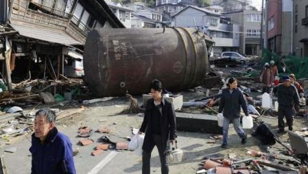 Supervivientes del tsunami caminan con envases de plástico y ollas para llevar agua potable en una una calle bloqueada por un tanque y otros escombros el pasado 14 de marzo. La foto superior muestra en la misma calle, el 3 de junio, sólo una casa dañada. la localidad es Kesennuma, Prefectura de Miyagi, al noreste de Japón.