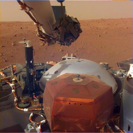 El sismómetro de InSight se encuentra por ahora encima de la sonda (bajo una cubierta protectora roja en la imagen), a la espera de que el brazo robótico (arriba) pueda lo pueda desplegar sobre la superficie de la Elysium Planitia en Marte