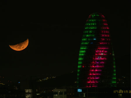 La Torre Glòries iluminada por la noche, junto a la luna creciente.