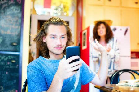 Male teenager ignoring scolding mom with mobile phone. He gestures her to leave him be as he smiles at his phone. Focus on the boy. (Male teenager ignoring scolding mom with mobile phone. He gestures her to leave him be as he smiles at his phone. Focu