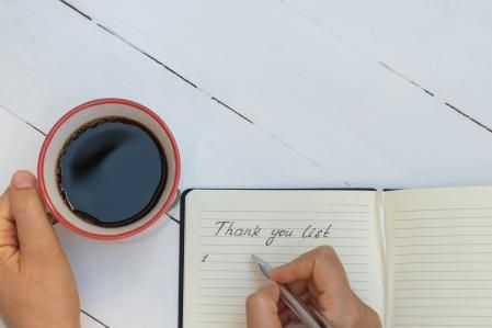 Top view of woman hand writing thank you list and holding cup of coffee on white wooden table. Motivation and inspiration concept (Top view of woman hand writing thank you list and holding cup of coffee on white wooden table. Motivation and inspiratio