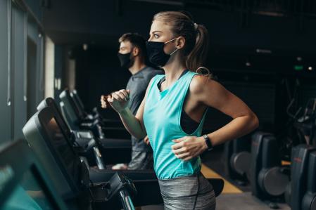 Young fit woman and man running on treadmill in modern fitness gym. They keeping distance and wearing protective face masks. Coronavirus world pandemic and sport theme. (Young fit woman and man running on treadmill in modern fitness gym. They keeping