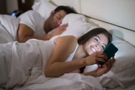 Woman texting or social networking on her smart phone in bed while her husband is sleeping