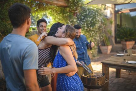 Young and mid adult Spanish female friends hugging and laughing while barbecuing at outdoor social gathering.