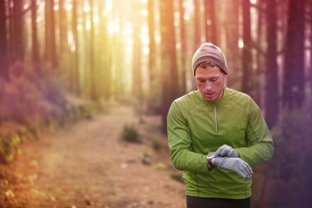 Trail running runner looking at heart rate monitor watch running in forest wearing warm jacket sportswear, hat and gloves. Male jogger running training in woods. (Trail running runner looking at heart rate monitor watch running in forest wearing warm