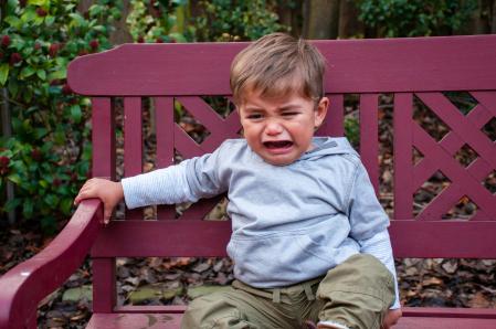 Upset child sitting on bench crying with greenery in the background.