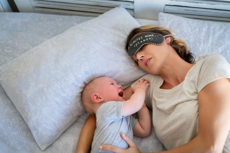 Tired mother with eye band trying to sleep while little toddler crying on bed. Angry baby boy crying and trying to wake mother. Stressed mother trying to take a nap with her child.