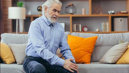 Un hombre mayor sentado en el sofá de su casa tiene dolor en la articulación de la rodilla