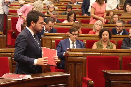 El president de la Generalitat, Pere Aragonès, dret i amb una carpeta a la mà amb el líder del PSC, Salvador Illa, de fons durant el ple del Parlament.