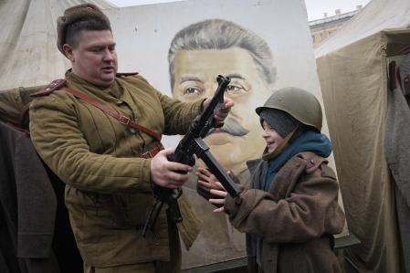 A member of a historic club wearing a World War II Soviet Army's uniform shows a gun to a boy near a portrait of Soviet dictator Josef Stalin at a military-historical exhibition at the Dvortsovaya (Palace) Square, in St. Petersburg, Russia, Saturday, Jan. 27, 2024. The exhibition marked the 80th anniversary of the battle that lifted the Siege of Leningrad. The Nazi siege of Leningrad, now named St. Petersburg, was fully lifted by the Red Army on Jan. 27, 1944. More than 1 million people died mainly from starvation during the nearly900-day siege. (AP Photo/Dmitri Lovetsky)