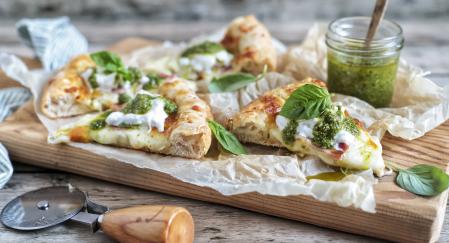 Trozos de pizza sobre una tabla de madera con un cortador y un bote de salsa pesto