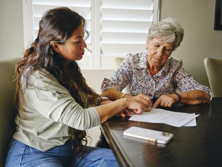 A Hispanic woman, helping a senior aged woman prepare financial documents.