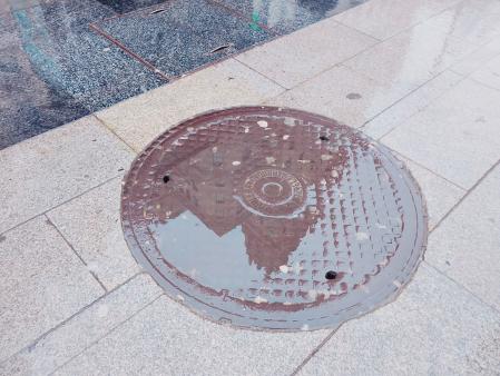 En la tapa de una alcantarilla se observa reflejado el edificio de la Telefónica.