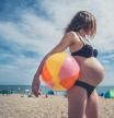 A young pregnant woman is standing on the beach with an inflatable beach ball