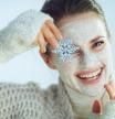 Portrait of smiling modern middle age woman in roll neck sweater and cardigan with white facial mask showing snowflake on winter light blue background. (Portrait of smiling modern middle age woman in roll neck sweater and cardigan with white facial ma
