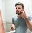 Young man taking care of his skin. Man applying skincare facial treatment cream on face in the bathroom. Handsome guy applying moisturizer and looking at himself while standing in front of the mirror.