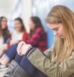 A high school girl sits alone by her locker, her head hung low in sadness, as a group of three friends in the background point at her and laugh.